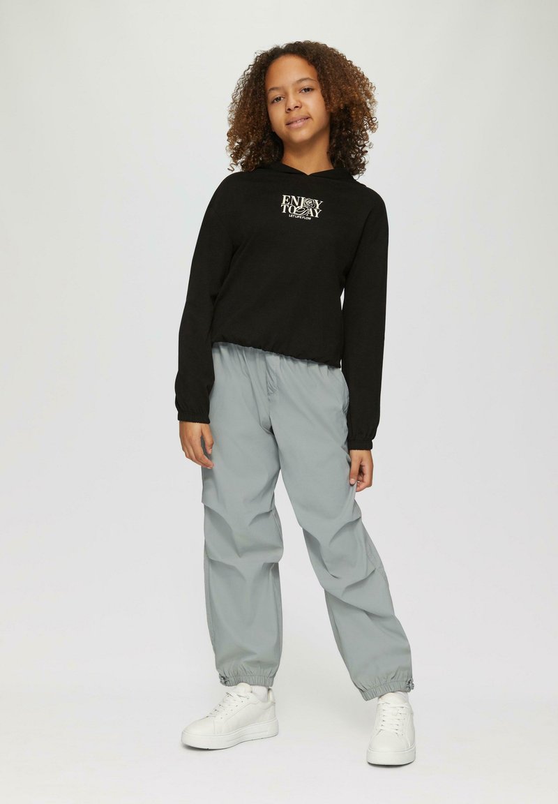 Felpa nera corta con stampa "Goditi Oggi", abbinata a pantaloni grigi chiari con polsini elastici e sneakers bianche. Abbigliamento sportivo semplice e casual.