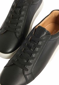 Para czarnych skórzanych sneakersów z czarnymi sznurówkami i białymi podeszwami, z widoczną teksturowaną powierzchnią i brązowym wewnętrznym wykończeniem.
