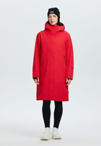 Giacca impermeabile lunga rossa con colletto alto e cappuccio, caratterizzata da una chiusura a zip e tasche laterali, abbinata a leggings neri e scarpe.