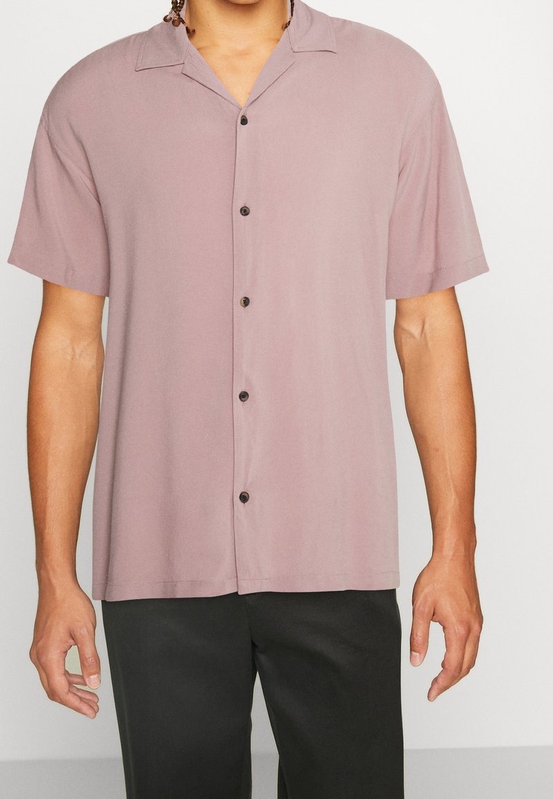Homme portant une chemise boutonnée rose poussiéreux à manches courtes avec un pantalon noir devant un fond clair uni.