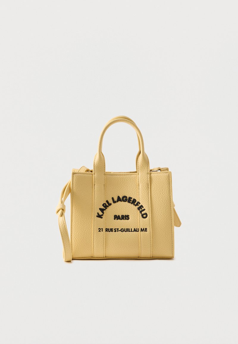 Μικρή κίτρινη υφασμάτινη τσάντα με δύο χερούλια, αφαιρούμενο λουρί και μαύρο κείμενο "Karl Lagerfeld Paris 21 Rue St-Guillaume" στο μπροστινό μέρος.