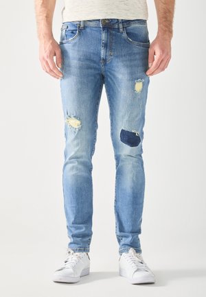 Homme portant un jean bleu clair usé avec des patchs et des baskets blanches, debout devant un fond blanc uni.
