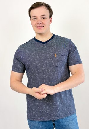 Jeune homme aux cheveux bruns courts, portant un t-shirt rayé bleu marine et un jean, souriant et joignant ses mains devant un fond uni.