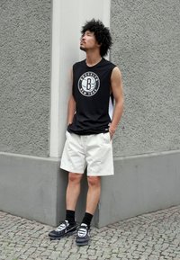 Camiseta negra sin mangas con el logotipo "Brooklyn New York", pantalones cortos blancos, calcetines negros y zapatillas negras con acentos claros, contra un fondo gris texturizado.
