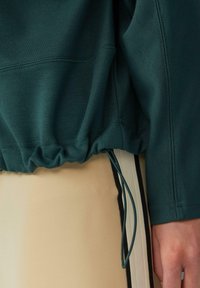 Primo piano di una mano appoggiata vicino all'orlo di un hoodie verde con coulisse e pantaloni beige con strisce laterali nere e bianche.
