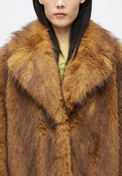 Žena v hrubej hnedej kožuchu cez svetlozelený top, ktorý odhaľuje golier a hornú časť tela, s rovnými čiernymi vlasmi.