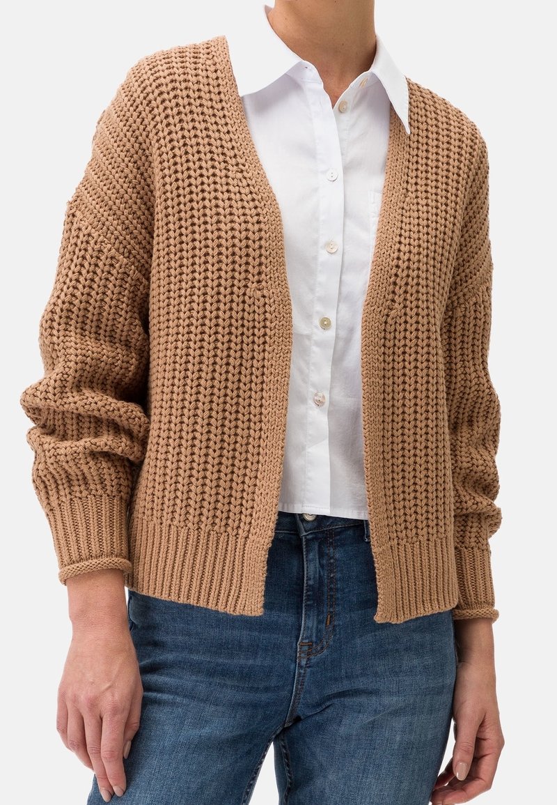 Brun stickad cardigan med överdimensionerade ärmar, öppen fram, ribbade mudd och fåll, som bärs över en vit skjorta med krage och jeans.