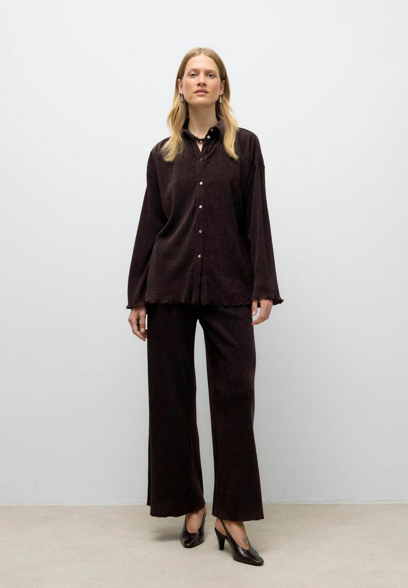 Vrouw die tegen een effen achtergrond staat, gekleed in een donkerbruine corduroy overhemd met knopen, bijpassende wijde broek en zwarte hakken.