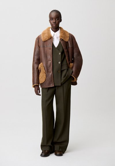 Chaqueta de cuero marrón con un cuello de borreguito color beige, combinada con un traje verde a medida y zapatos marrones, que presenta botones y bolsillos prominentes.