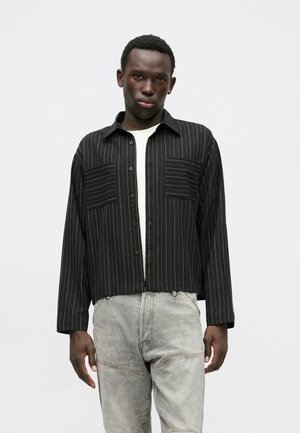 Mustriga must-valge särg, millel on kaks esitaskut, lühendatud disain ja pikad varrukad, kombinatsioonis heledate hallide pükstega.