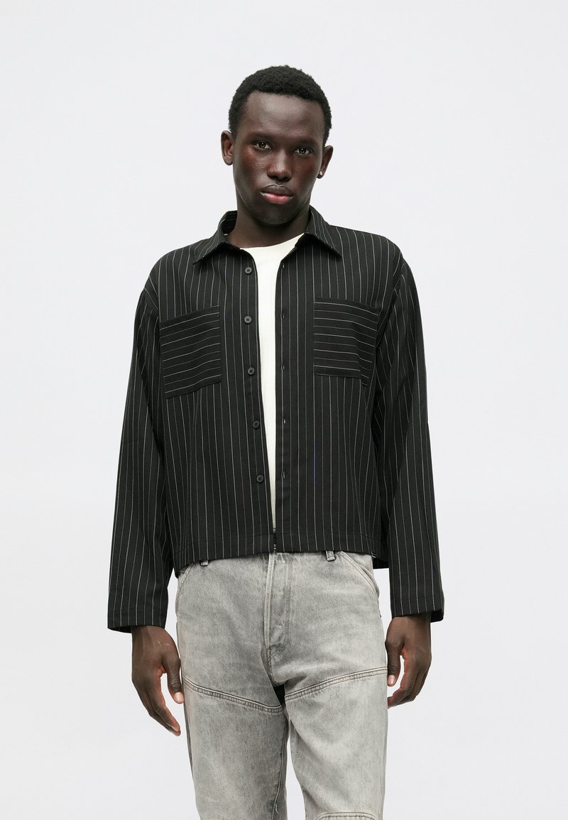 Camisa preta de riscas com dois bolsos frontais, design cropped e mangas longas, combinada com calças cinzas claras.