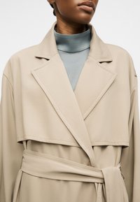 Gros plan sur une personne portant un trench-coat beige ceinturé par-dessus un haut bleu clair à col montant, montrant les détails du col et du haut de la poitrine.