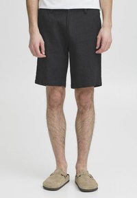 Fekete len nadrág rövidnadrág egyenes szabással, két elülső zsebbel és texturált anyaggal. Bézs slip-on cipővel viselve.