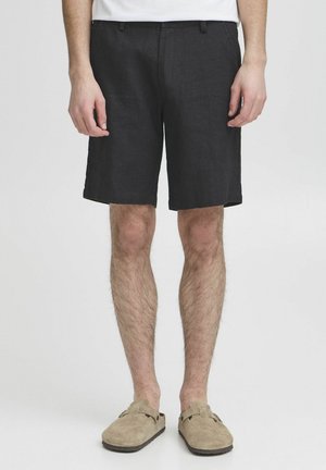 Shorts en lin noirs à coupe droite, avec deux poches avant et un tissu texturé. Portés avec des chaussures à enfiler beiges.