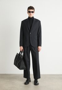 Completo nero con un blazer aderente e pantaloni a gamba ampia, abbinato a un dolcevita. Gli accessori includono occhiali da sole neri e una borsa strutturata.