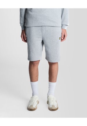Mand iført lysegrå sweatshirt og matchende shorts med hvide sokker og beige hvide sneakers, stående mod en ensfarvet lys baggrund.
