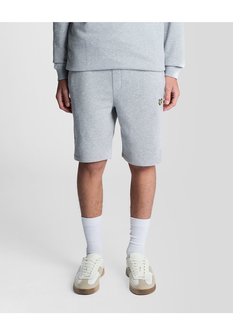 Homme portant un sweat-shirt gris clair et un short assorti avec des chaussettes blanches et des baskets beiges et blanches, debout devant un fond clair uni.