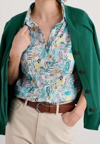 Cardigan vert porté sur une chemise à motifs floraux multicolores. La chemise présente des dessins bleus, jaunes et violets. Pantalon beige clair et ceinture marron.