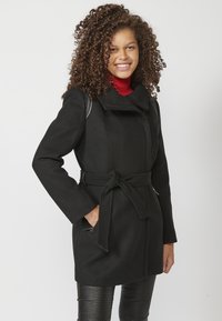 Cappotto di lana nera con vita abbottonata, colletto alto, tasche con zip e maniche lunghe. Indossato sopra un maglione aderente rosso e pantaloni texturizzati.