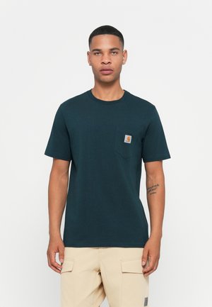 Man wearing dark green Carhartt pocket t-shirt and light beige cargo pants, standing against a plain white background.
