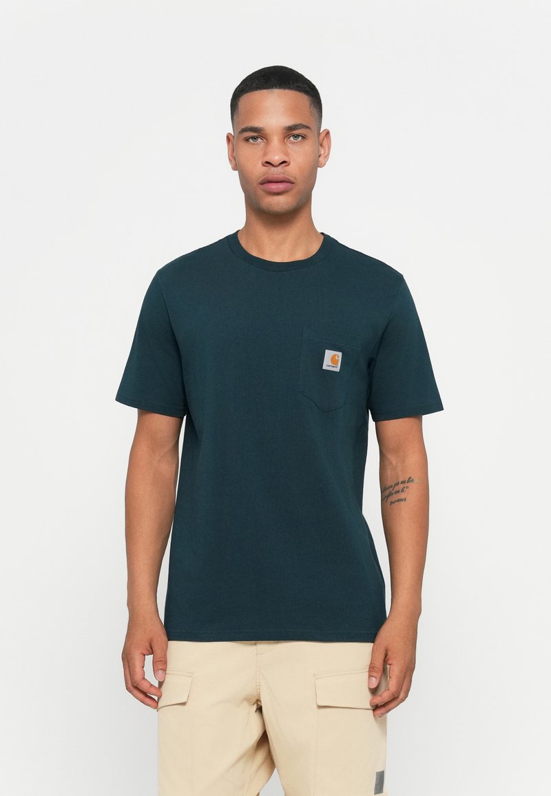 Man in een donkergroene Carhartt pocket t-shirt en lichtbeige cargobroek, staande voor een effen witte achtergrond.
