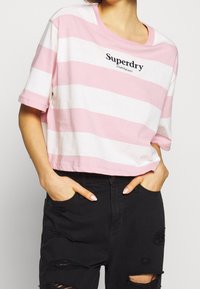 Světle růžové a bílé pruhované zkrácené tričko s kulatým výstřihem. Obsahuje logo "Superdry" v černé barvě a volné krátké rukávy.