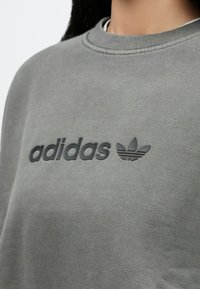 Γκρι φούτερ από μαλακό ύφασμα, με ανάγλυφο μαύρο λογότυπο "adidas" και σχέδιο με τρεις λωρίδες σε σχήμα φύλλου στο μπροστινό μέρος. Κλασική στρογγυλή λαιμόκοψη.