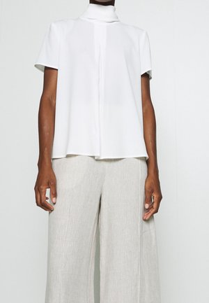 Persona con blusa blanca de manga corta y cuello alto, y pantalones holgados de lino beige, sobre un fondo claro y liso.