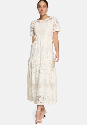 Kobieta w białej, koronkowej sukience maxi z krótkimi rękawami i kwiatowym wzorem, z warstwowym dołem, w połączeniu z białymi sandałkami na obcasie.
