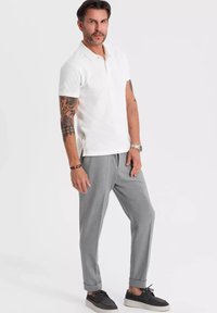 Vit polotröja, grå tapered byxor, svarta sneakers och en klocka. Har tatueringar på armarna och uppvikta byxor. Enkel design, avslappnad stil.