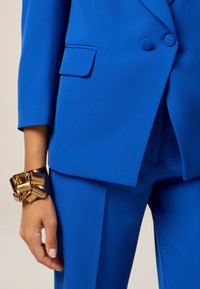 Cobalt blue tailored suit with structured blazer, featuring a notch lapel and one patch pocket. The wrist adorned with a chunky gold bracelet.