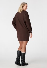 Bruine jurk met lange mouwen en een ontspannen silhouet, gemaakt van gladde stof. Gecombineerd met kniehoge zwarte laarzen met metalen accenten.