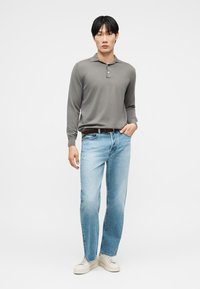 Άνδρας που φορά γκρι πόλο με μακριά μανίκια, ανοιχτό μπλε τζιν, καφέ ζώνη και λευκά αθλητικά παπούτσια, στέκεται μπροστά σε απλό φόντο.