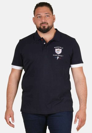 Homme portant un polo bleu marine avec le logo Ruckfield et le texte "Allez Galle !", debout en posture détendue devant un fond blanc.