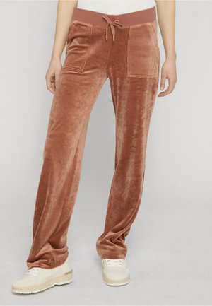 Person, die rostfarbene Velourhosen mit aufgesetzten Fronttaschen und Kordelzug an der Taille trägt, kombiniert mit weißen Sneakern vor grauem Hintergrund.