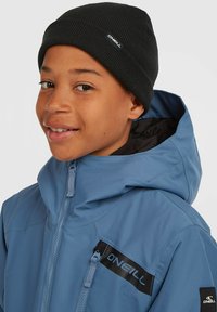 Blå zip-jacka med huva, med en svart accent på bröstet som visar märket "ONeill." Svart ribbad mössa på huvudet.