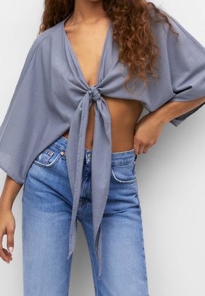 Kobieta w luźnym szarym przywiązanym krótkim topie z szerokimi rękawami i jasnoniebieskich jeansach z wysokim stanem, pozująca z dłonią na biodrze.