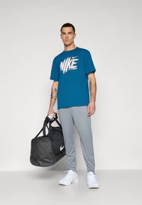 Nike sportswear-outfit med en teal t-shirt med hvidt logo, grå bukser og en sort træningstaske. Skoene er hvide med teksturerede detaljer.