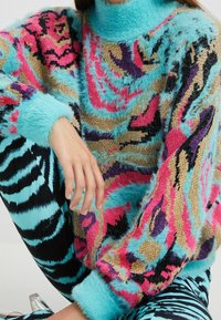 Farverig sweater med pelsagtig tekstur, med et mønster af pink, lilla og guld mod en turkis baggrund. Ribstrikkede manchetter og halsudskæring.