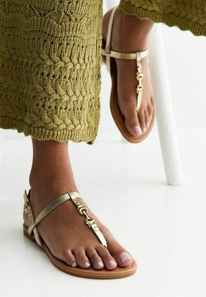 Pies con sandalias metálicas doradas de tiras en T y pantalones trenzados de punto verde oliva, apoyando un pie en la pata blanca de una silla.