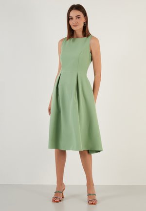 Light green sleeveless dress with a fitted bodice and pleated skirt; smooth fabric, mid-calf length, paired with strappy open-toed heels.