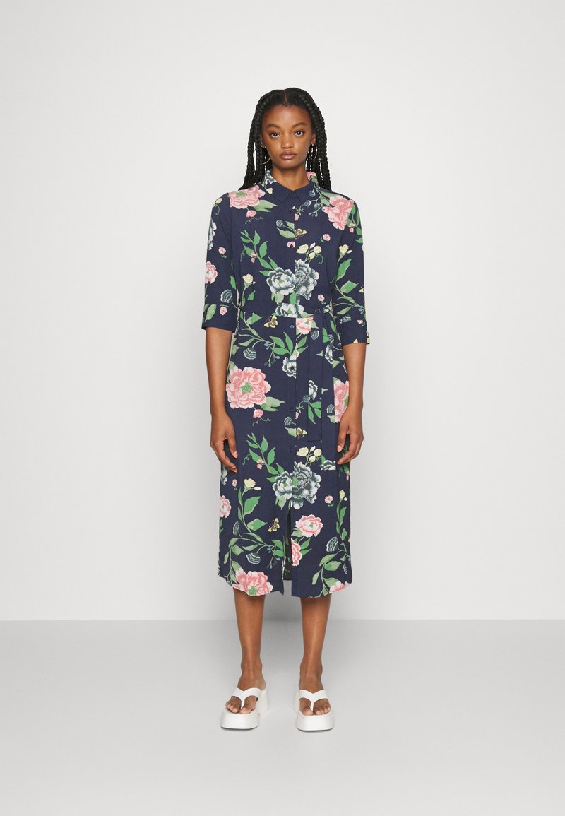 Vestido floral em azul-marinho com mangas metade, cintura cingida e corte até ao joelho; apresenta padrões de flores em rosa e verde. Combinado com sandálias de plataforma brancas.