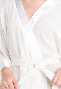 Ropa blanca texturizada con un escote de satén y cinturón. Presenta un diseño envolvente con mangas largas y un brillo sutil.