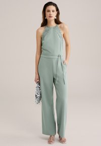 Mintgrön jumpsuit med halterneck, spetsdetaljer, vida ben och knytmidja. Modellen håller en silverfärgad metallisk clutch.