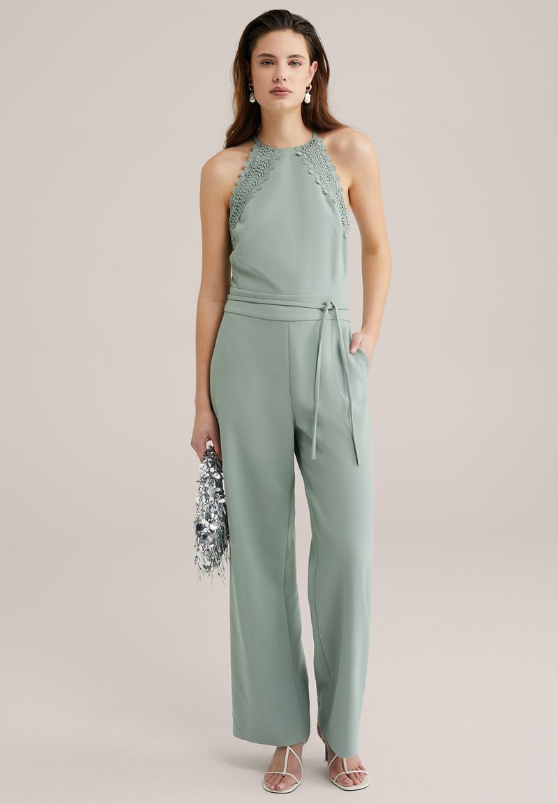Mintgrön jumpsuit med halterneck, spetsdetaljer, vida ben och knytmidja. Modellen håller en silverfärgad metallisk clutch.