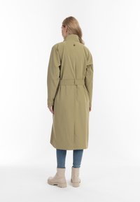Olijfgroene, lange trenchcoat met een hoge kraag, een ceintuur in de taille en een achterkantventilatie, gecombineerd met lichtgekleurde enkellaarsjes en blauwe spijkerbroeken.