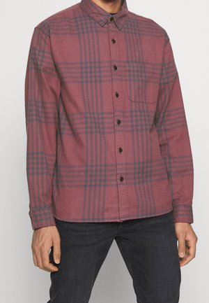 Mand iført en langærmet bordeauxrød ternet skjorte med sorte knapper og en brystlomme foran, kombineret med sorte bukser.