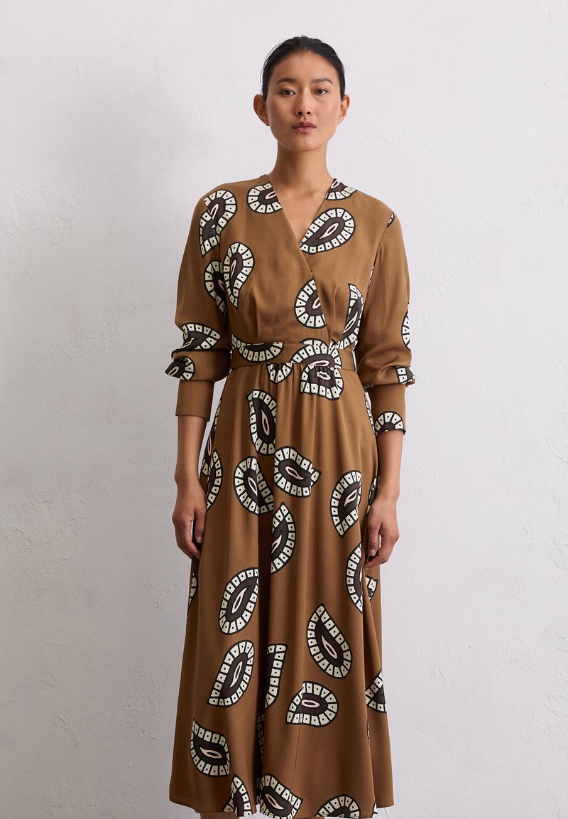 Braunes Wickelkleid mit langen Ärmeln, das schwarz-weiße Kreis-Muster aufweist. Glatte Textur mit betontem Taillenschnitt, der bis zur Mitte der Wade reicht.