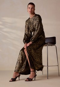 Maxikleid mit Leopardenmuster in Gold und Schwarz, langen Puffärmeln und Schlitz. Kombiniert mit schwarzen Sandalen mit Absatz, die eine Schleife und Nietenverzierung aufweisen.