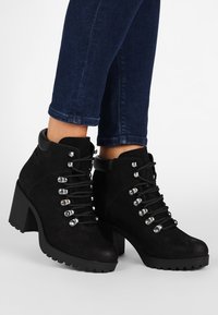 Zwarte suède enkellaarsjes met een stevige hak, voorzien van metalen oogjes, zwarte veters en een textuurrijke zool voor extra grip.